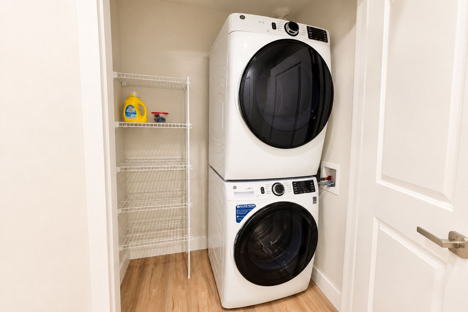 A laundry room with two stacked washing machines, a shelf with cleaning supplies, and a door.