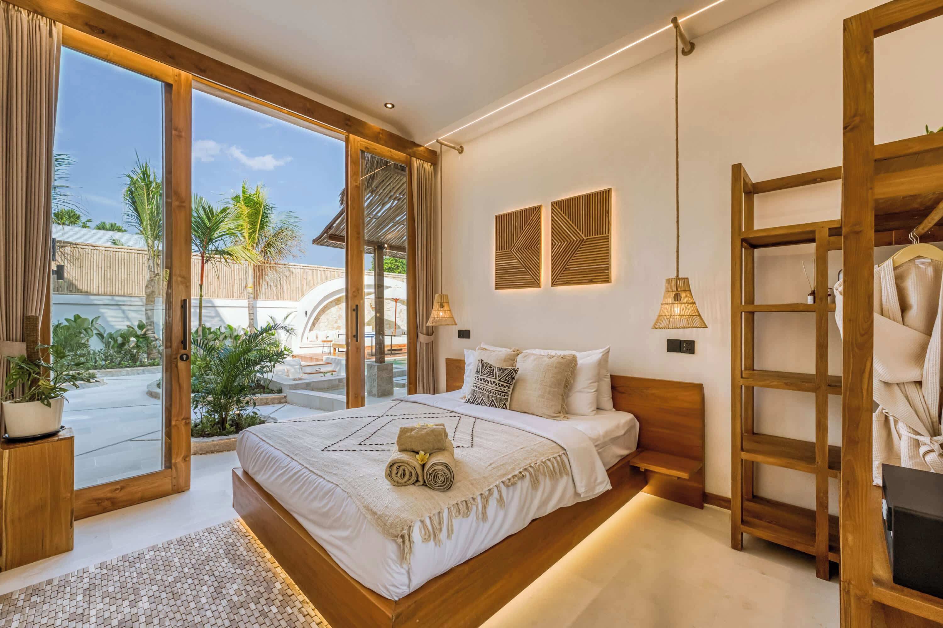 Une chambre avec un grand lit, une tête de lit en bois, un balcon avec vue sur les palmiers et un fauteuil suspendu.