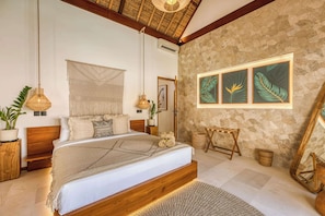 Ein geräumiges Schlafzimmer mit einem großen Bett, einer Steinwand und gerahmten Kunstwerken mit tropischen Blättern.