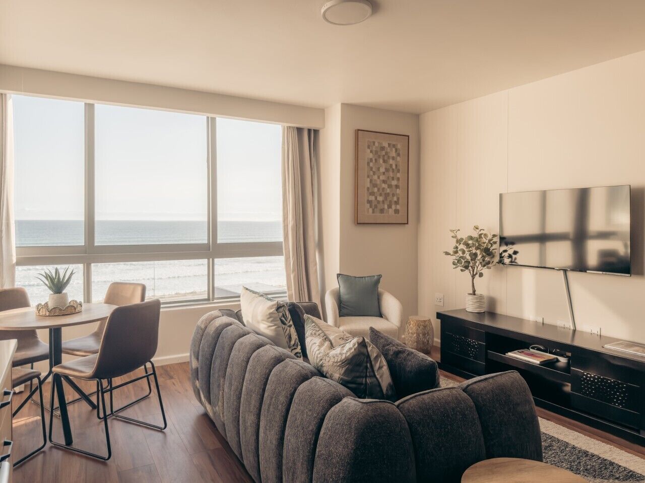 A modern living room with a large sofa, a dining area, and a TV mounted on the wall.