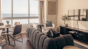 A modern living room with a large sofa, a dining area, and a TV mounted on the wall. - Infinity 2BR Seaside Stay - Steps from Blouberg Beach (Cape Town)
