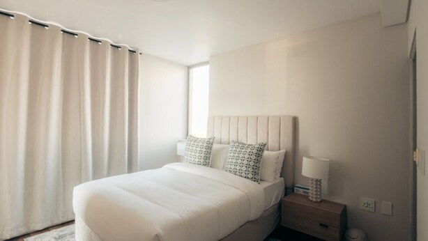 A modern bedroom with a large bed, bedside tables, and a nightstand. - Infinity 2BR Seaside Stay - Steps from Blouberg Beach (Cape Town)