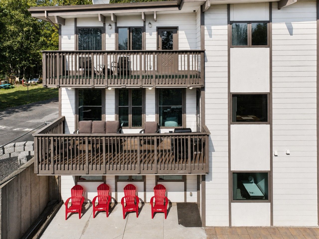 A multi-story building with a balcony on the ground floor, featuring outdoor seating and red chairs.