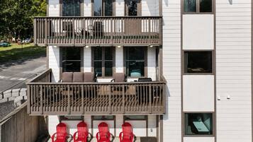 A multi-story building with a balcony on the ground floor, featuring outdoor seating and red chairs.