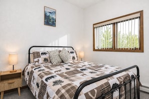 A bedroom with a bed, two bedside tables, a window with blinds, and a painting on the wall. - Aurora Skyline Lodge (North Pole)