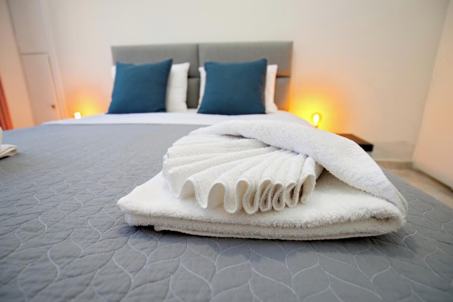 A neatly made bed with a folded white towel in the center, a grey quilted bedspread, and two blue pillows on a headboard.