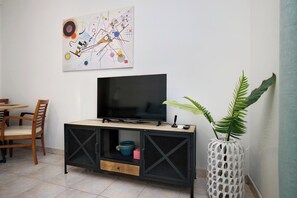 Uma sala de estar com uma TV em um móvel preto, um quadro na parede e um vaso com planta.