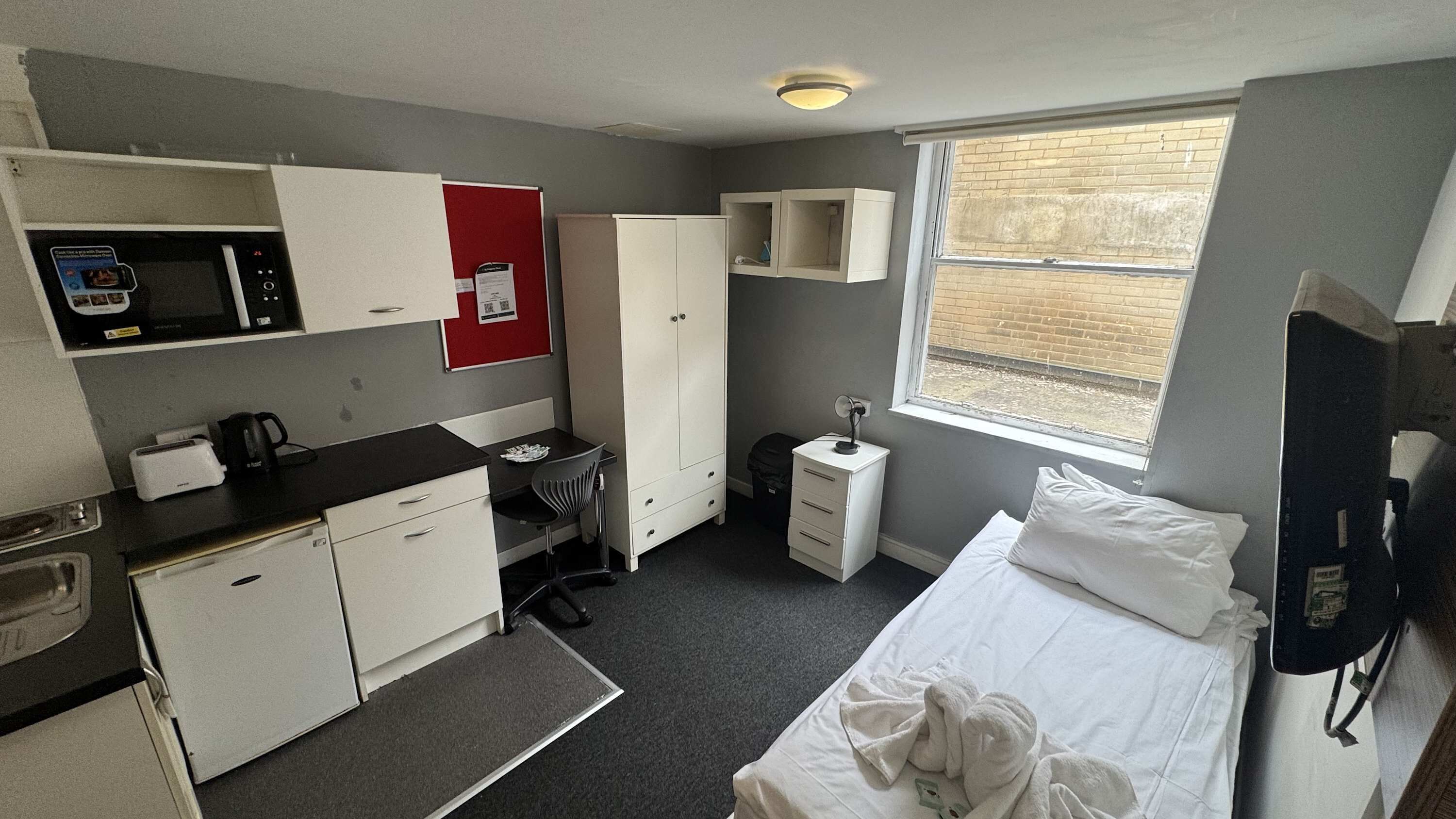 Ein kleines Hotelzimmer mit Kochnische, einem Bett, einem Nachttisch, einem Fenster und einem Kleiderschrank.