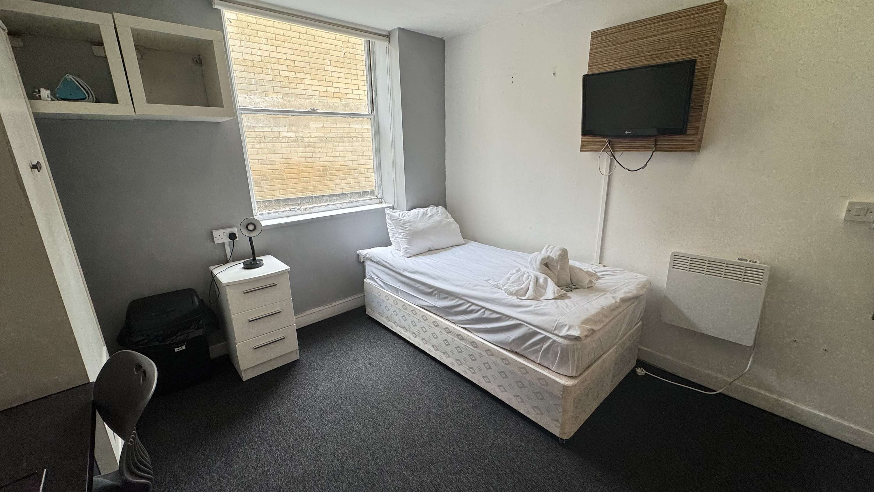 A small, neatly arranged bedroom with a single bed, a desk with a chair, a shelf unit, and a TV mounted on the wall.