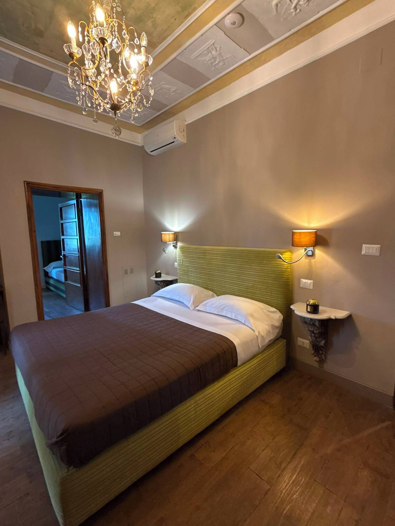 Een hotelkamer met een groot bed, een kroonluchter, wandlampen en een deur die naar een andere kamer leidt.