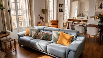 A modern living room with a grey sofa, wooden flooring, and a dining area with a white table and chairs.