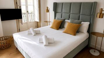 A neatly made bed with white linens and two pillows, a television mounted on the wall, and a window with curtains.