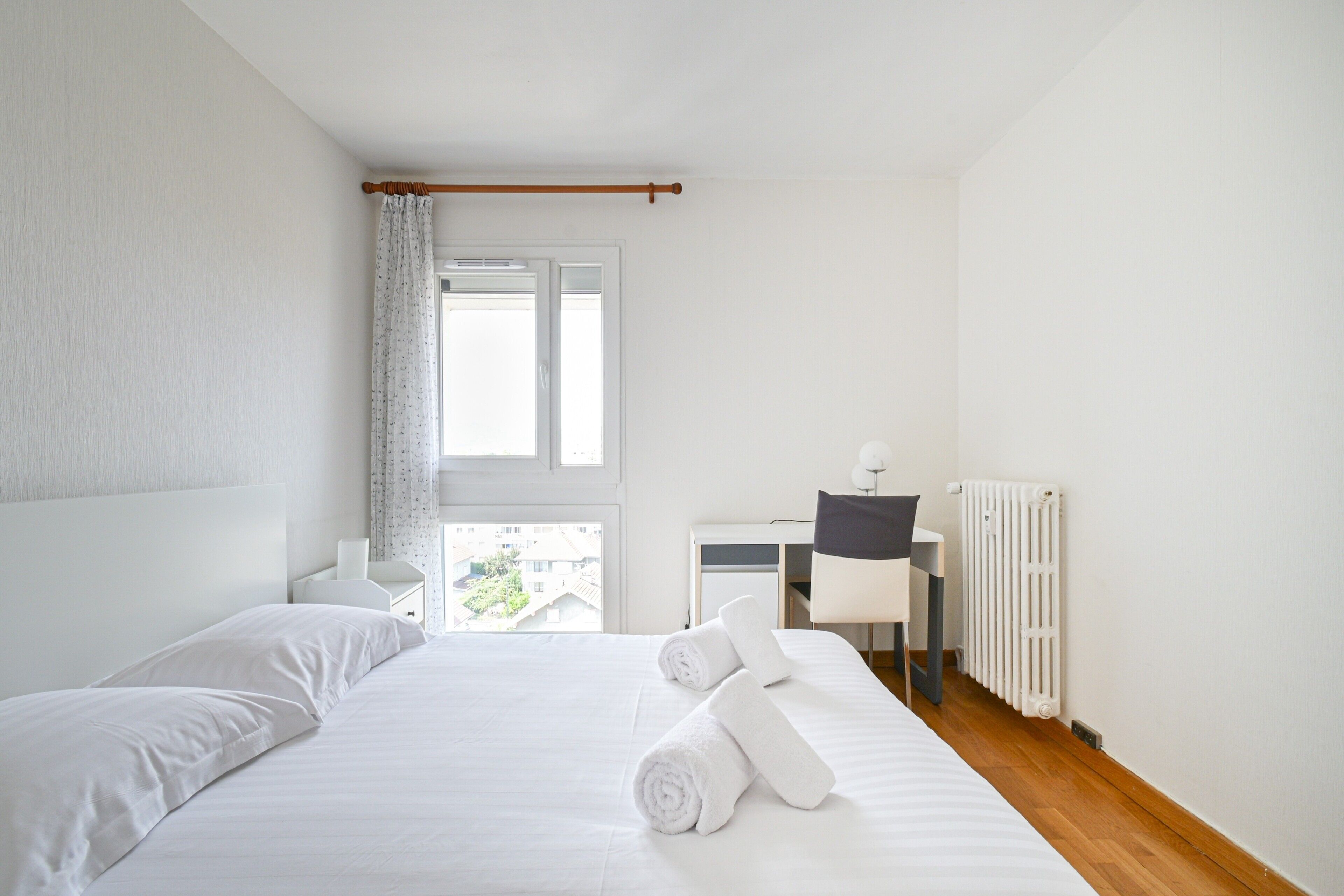 A hotel room with a bed, a desk, a chair, a radiator, and a window with curtains.