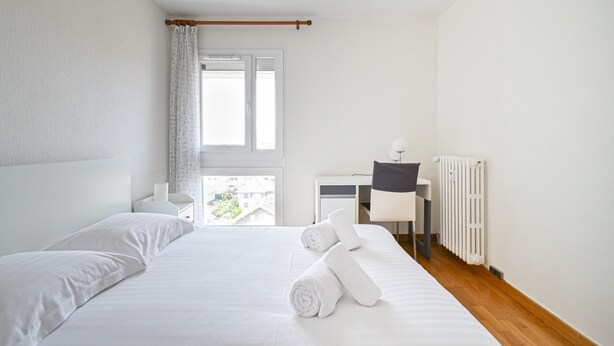 Ein Hotelzimmer mit Bett, Schreibtisch, Stuhl, Heizkörper und einem fenster mit Vorhängen.