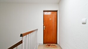 Ein schmaler Flur mit einer Holztür am Ende, einem weißen Geländer links und einer Fußmatte mit 'I ♥ Home' auf dem Boden.