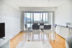 A modern living room with a dining area, large windows, and a view of buildings and mountains. - Spacious T4 Apartment with Mountain Views, Wi-Fi, and Parking – Sleeps 8 in Annecy (Annecy)