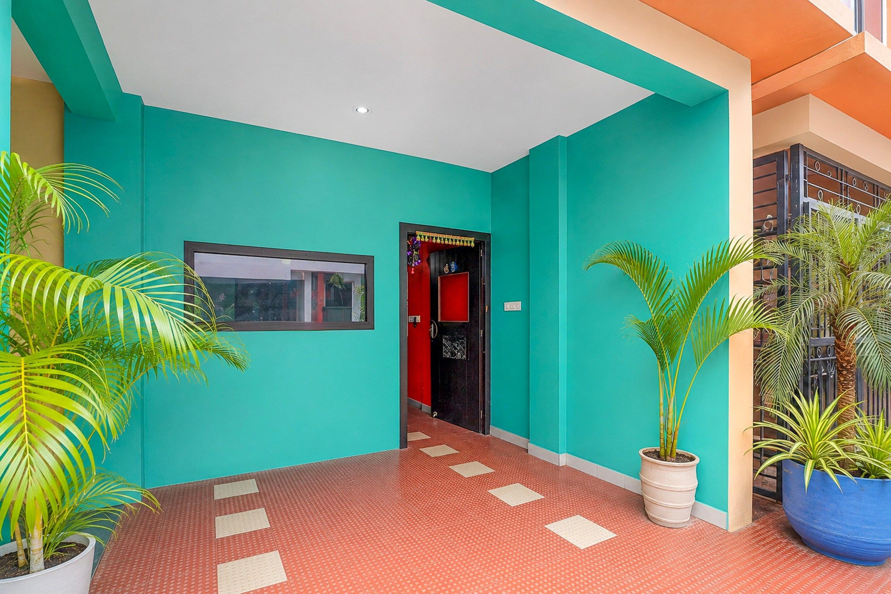 A hallway with a red tiled floor, a teal wall, and a black door with a red frame. There are potted plants on either side of the hallway.