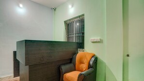 A reception area with a dark reception desk, a single leather chair, a window with metal bars, and a light switch on the wall. - Classic Avisi Residency (Bhubaneshwar)