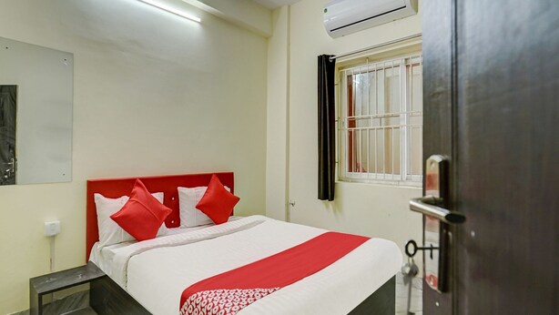 A hotel room with a bed, a nightstand, a window with bars, an air conditioning unit, and a door with a key lock. - Classic Avisi Residency (Bhubaneshwar)