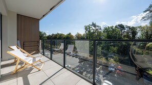 A balcony with a glass railing, wooden chairs, and a view of a parking area and trees. - Apartamenty Sun & Snow Koniecznego (Dziwnów)
