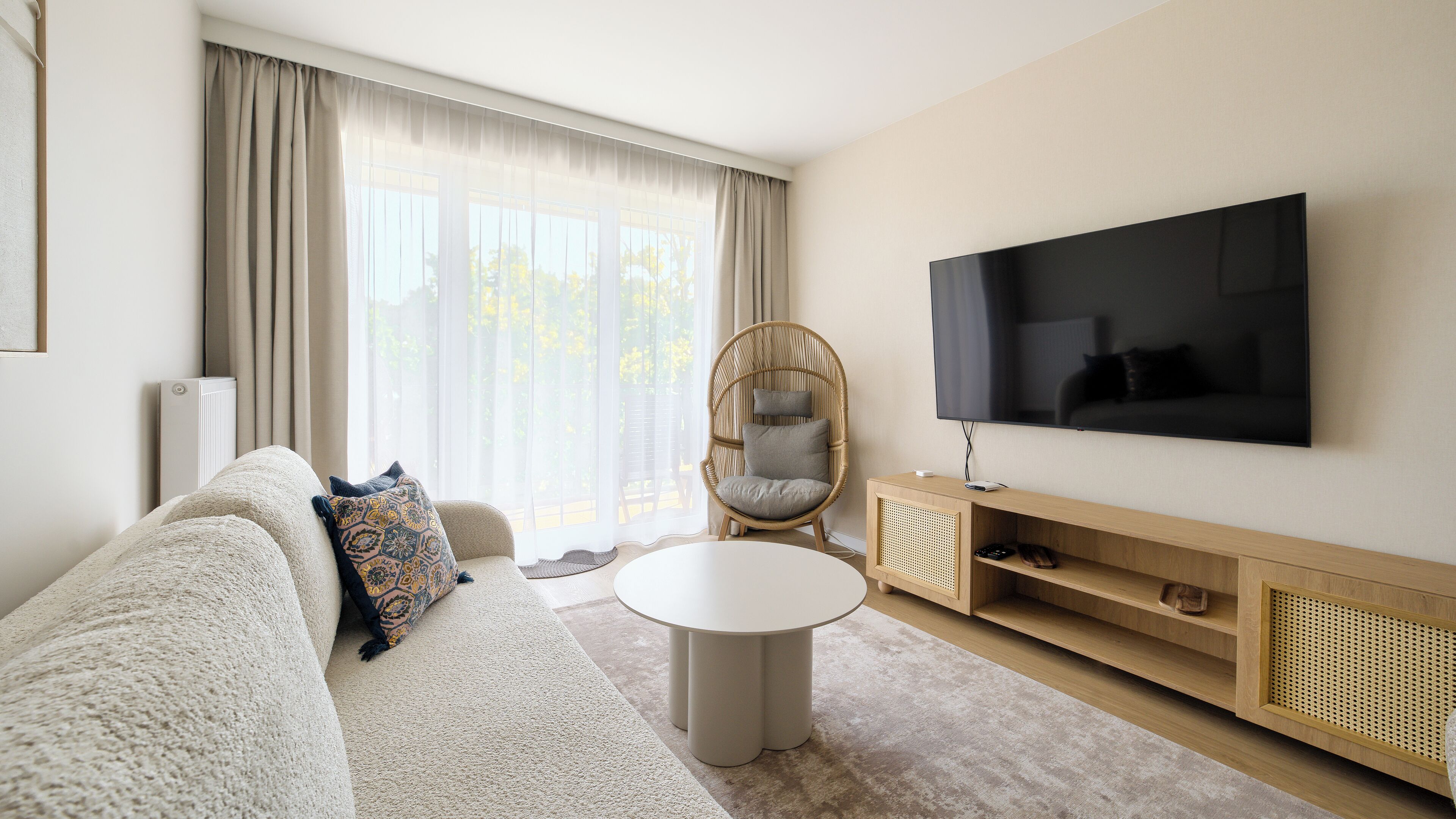 A modern living room with a sofa, a round coffee table, a wicker chair, a TV, and a large window with curtains.
