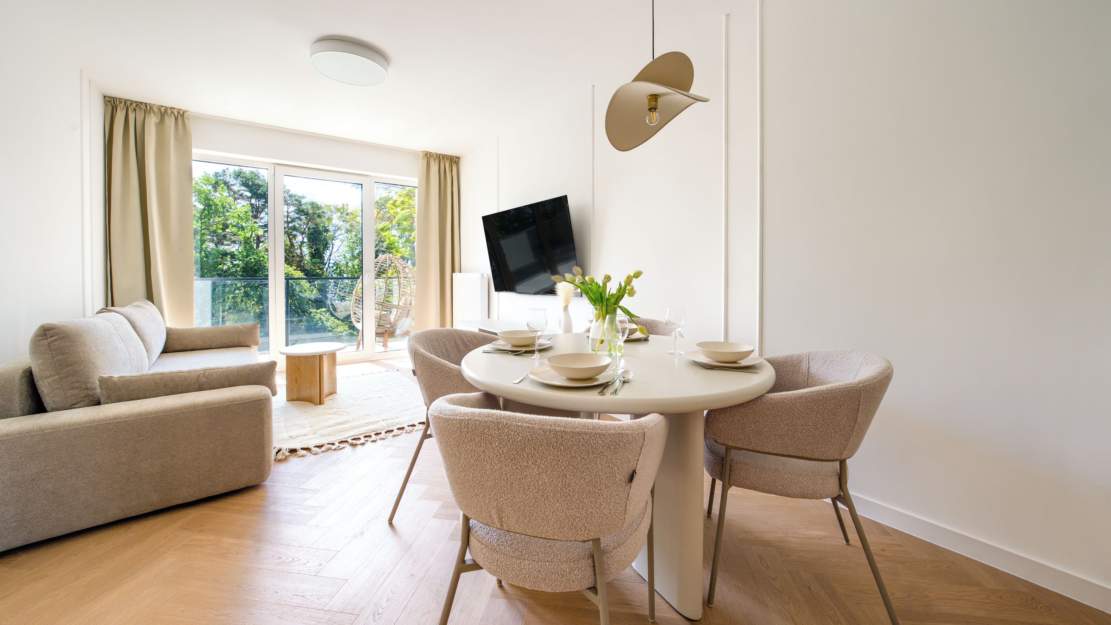 A modern living room with a round dining table, beige sofas, and a flat-screen TV mounted on the wall.