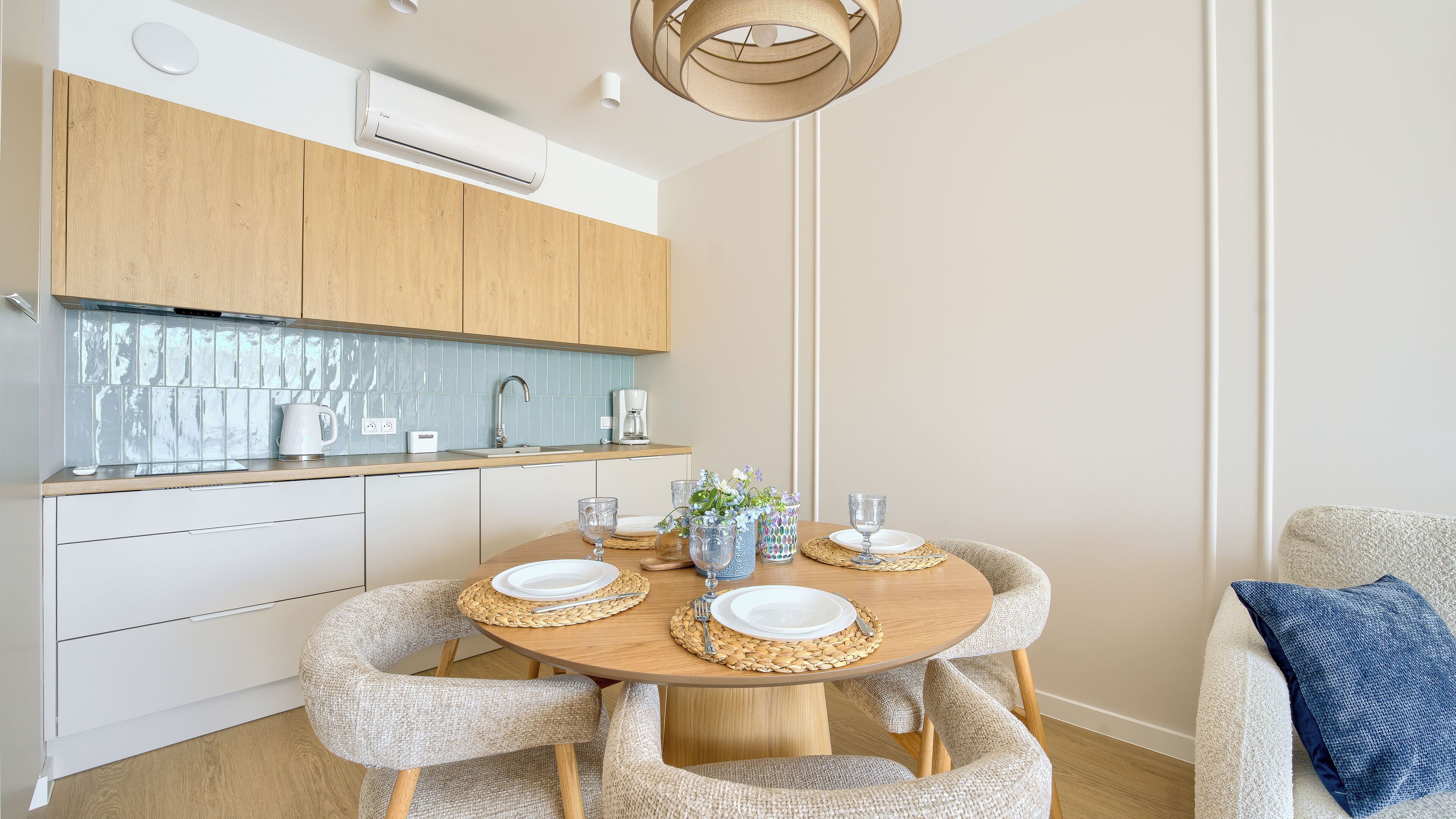 A modern kitchen with a dining area, featuring a round wooden table with white plates, woven placemats, and upholstered chairs.
