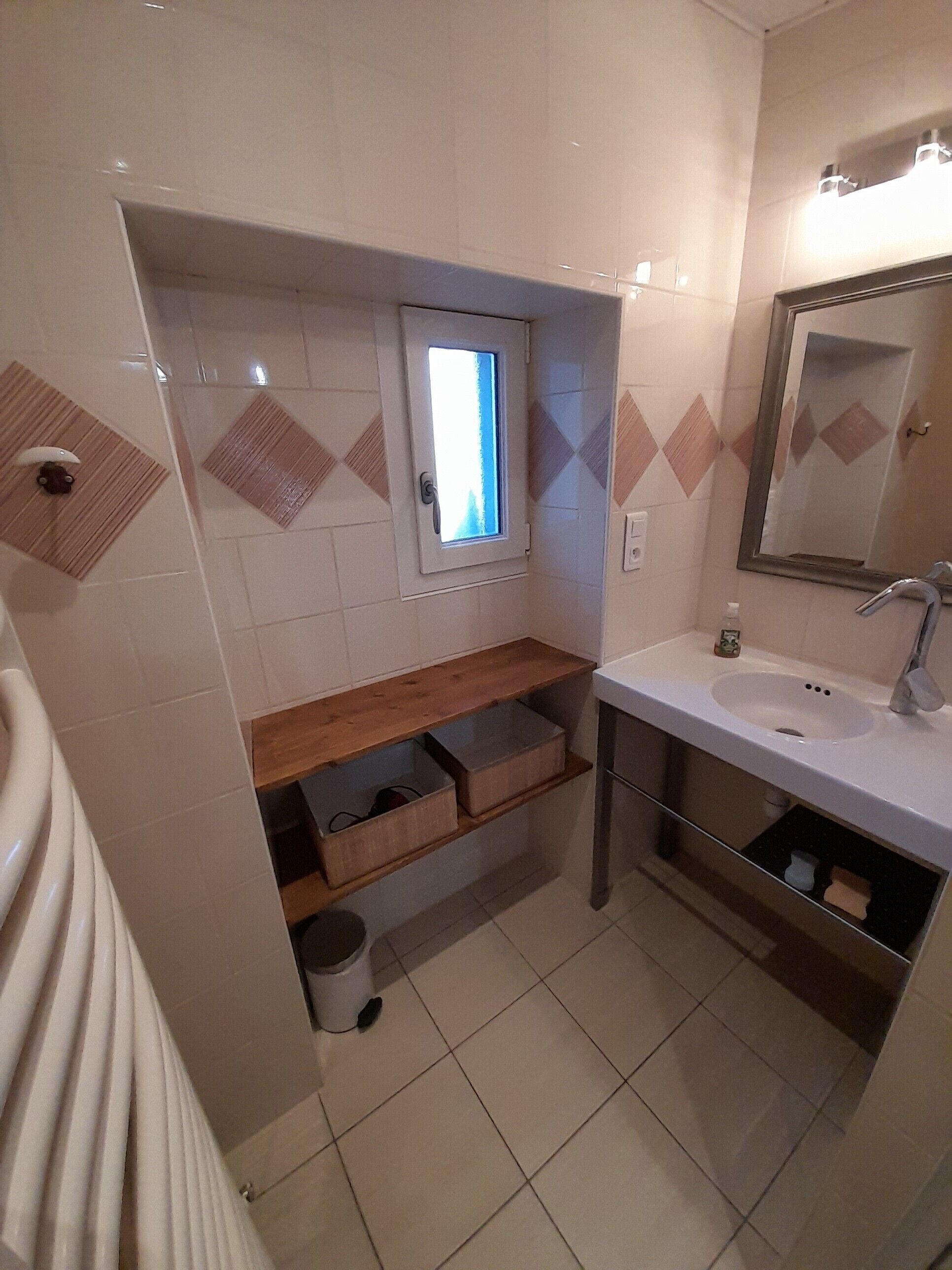 Ein Badezimmer mit Fenster, Waschbecken, Spiegel und Holzregal.