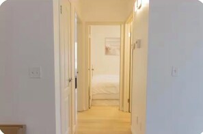 A narrow hallway leading to a bedroom with a bed, a door on the left, and a light switch on the wall.
