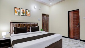 A hotel room with a bed, a nightstand, a lamp, a door, and three framed pictures on the wall. - Classic SGD Lawn And Hotel (Varanasi)