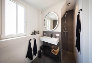 A modern bathroom with a large window, a round mirror, a white sink, a shower, and a small wicker basket. - Premium Outdoor 4p Villa in Thorn (Thorn)