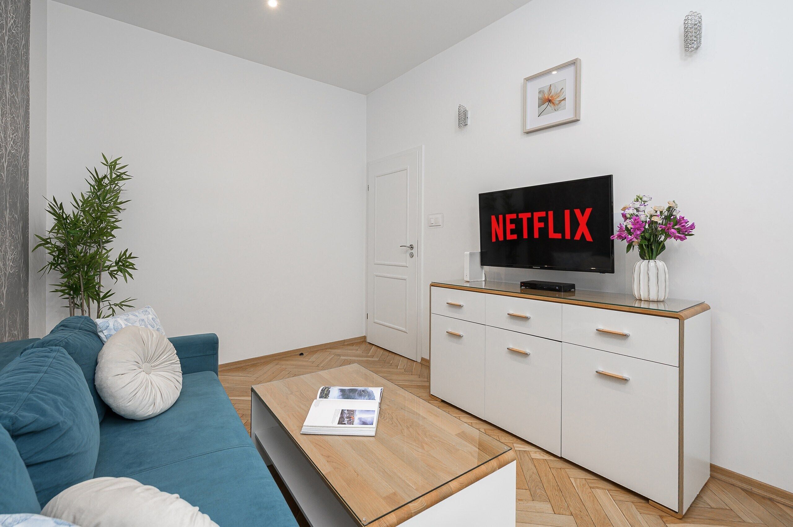 Un saló modern amb un sofà blau, una taula de centre amb una revista, un moble blanc amb una televisió que mostra 'NETFLIX' i una planta en test.