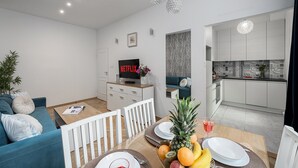 A modern living room with a sofa, a dining table set with plates and fruit, a TV, and a kitchen with white cabinets and patterned backsplash. - Lumina 3room Apart near Pałac Kultury (Warszawa)