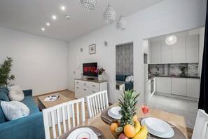 A modern living room with a sofa, a dining table set with plates and fruit, a TV, and a kitchen with white cabinets and patterned backsplash. - Lumina 3room Apart near Pałac Kultury (Warszawa)