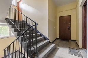 A staircase with a metal railing leading to a landing, a wooden door, and a window with a view of greenery. - Lumina 3room Apart near Pałac Kultury (Warszawa)