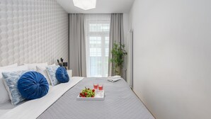 A modern bedroom with a bed, pillows, a bedside table, a window with curtains, and a tray with fruits and glasses. - Lumina 3room Apart near Pałac Kultury (Warszawa)