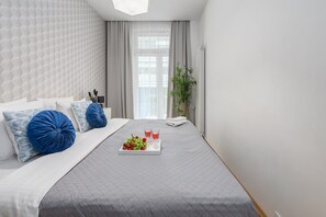 A modern bedroom with a bed, pillows, a bedside table, a window with curtains, and a tray with fruits and glasses. - Lumina 3room Apart near Pałac Kultury (Warszawa)