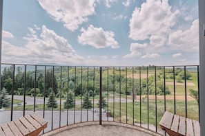 A balcony with a metal railing, wooden benches, and a view of a green landscape with trees and a road.