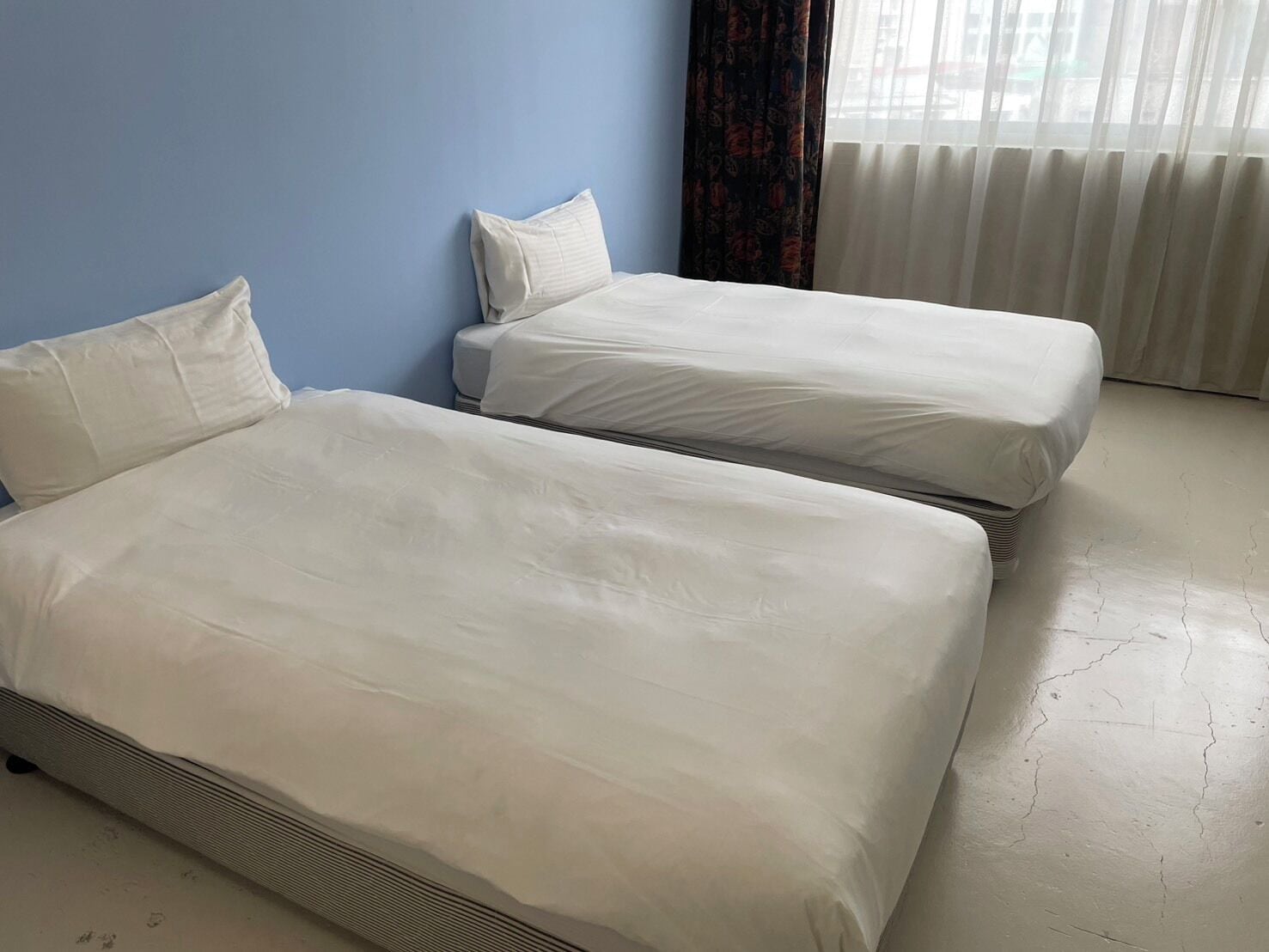 Ein Zimmer mit zwei Betten, einer blauen Wand und einem Vorhangfenster.