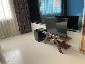 A modern living room with a flat-screen TV on a wooden stand, a small black speaker, and a window with curtains. - MUJU Hotel (Taipei)