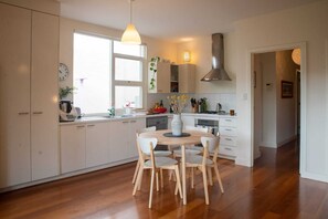 A modern kitchen with white cabinets, a wooden dining table, and a large window. - Cute & cosy family home - close to the city (Brompton)
