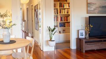 A living room with a wooden floor, a dining table with chairs, a bookshelf, a television, and a potted plant.