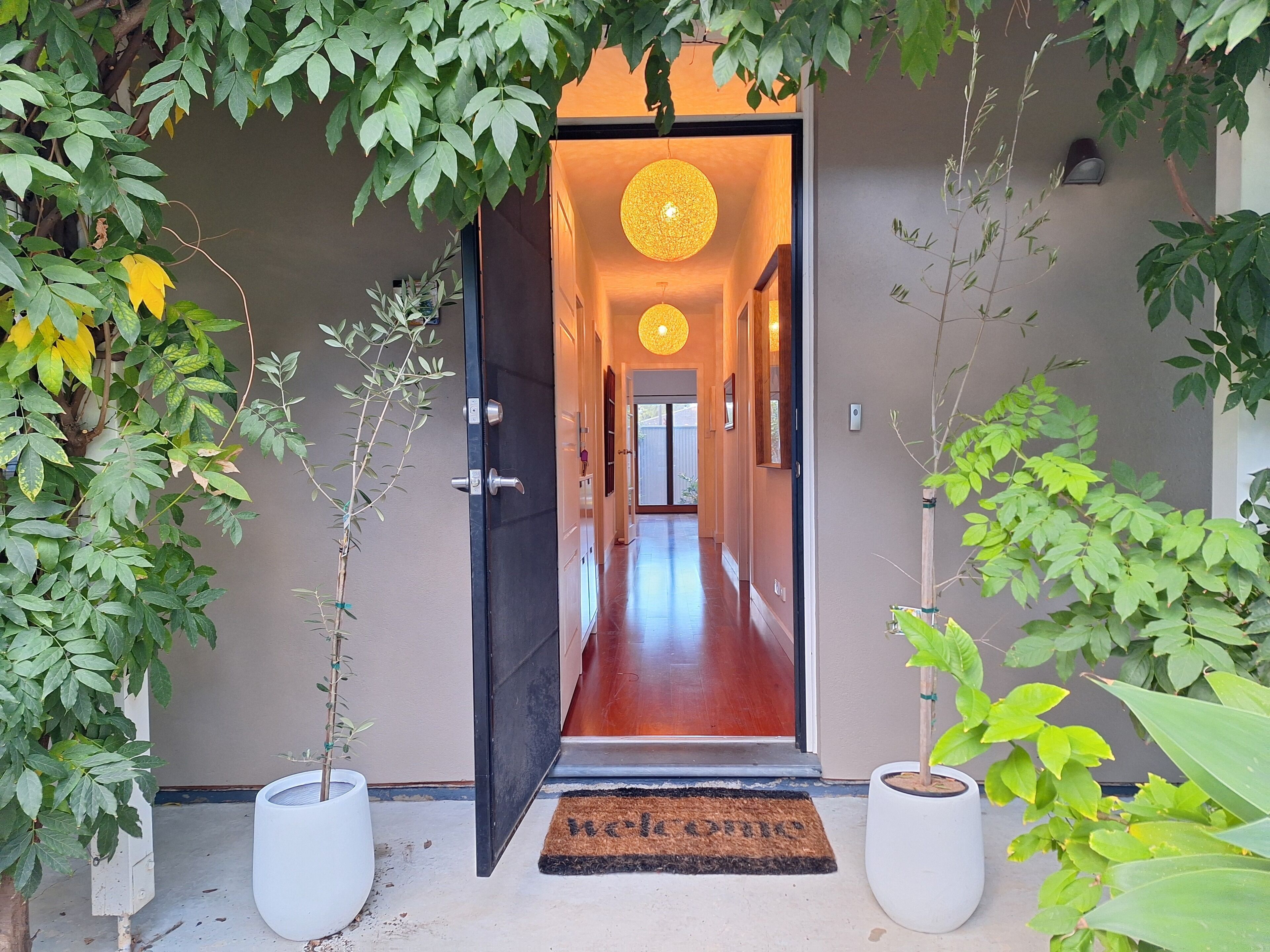 A hallway with a door leading to a room, potted plants on either side, and a doormat at the entrance.