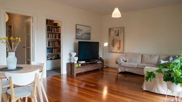 A living room with a wooden floor, a sofa, a television, a bookshelf, and a dining area with a table and chairs.