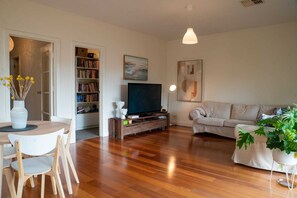 A living room with a wooden floor, a sofa, a television, a bookshelf, and a dining area with a table and chairs. - Cute & cosy family home - close to the city (Brompton)