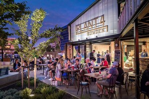 An outdoor seating area with tables and chairs, a building with a sign reading 'PLANTA BOWDEN,' and a person standing at a counter. - Cute & cosy family home - close to the city (Brompton)