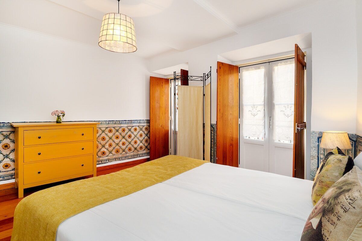 Une chambre à coucher avec un lit, une commode en bois, un luminaire suspendu et une fenêtre avec des rideaux.
