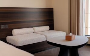 A hotel room with a dark wood headboard, a beige sofa, a round table, and a window with curtains.