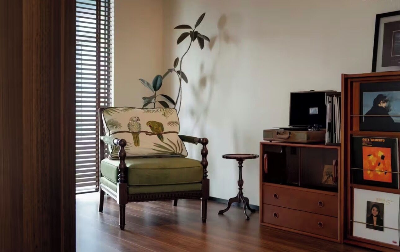 A room with a green armchair, a small wooden side table, a bookshelf, and a window with blinds.