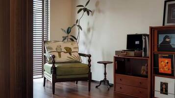 A room with a green armchair, a small wooden side table, a bookshelf, and a window with blinds.